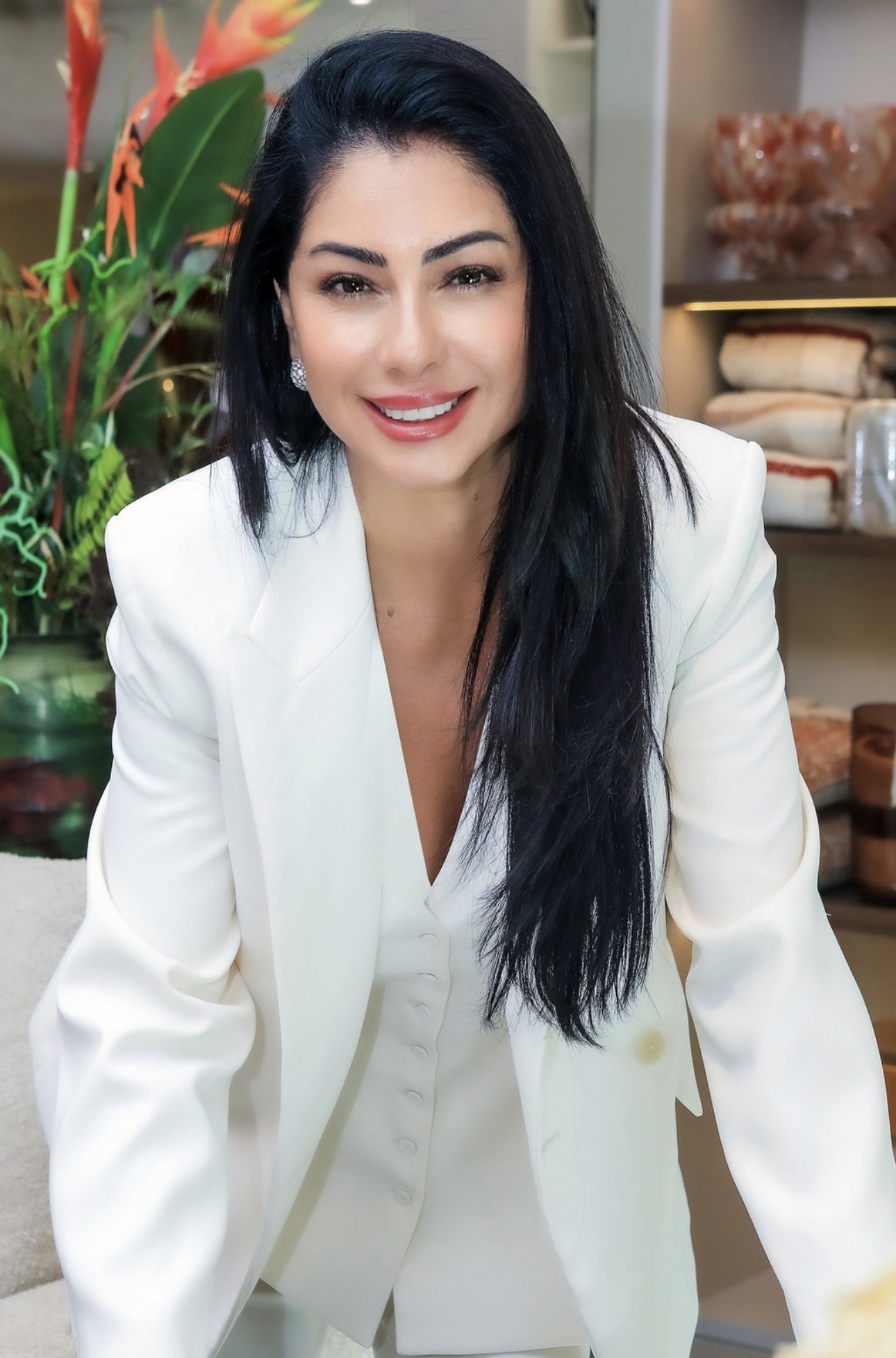 Andreia Camargo con blazer blanco sonriendo junto a una planta de flores naranjas